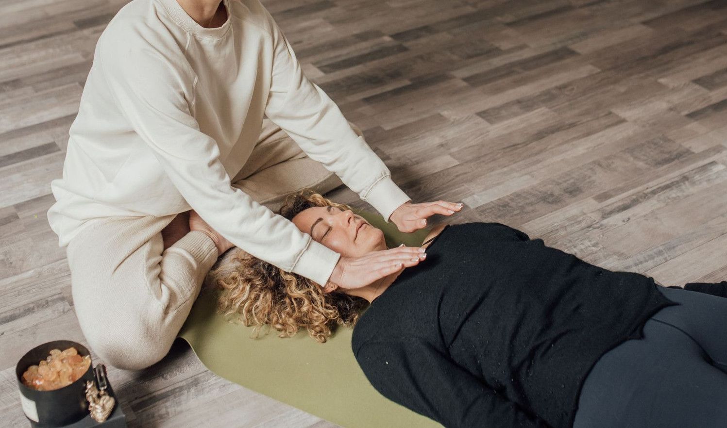 Woman practicing Reiki healing therapy on another woman lying indoors on a yoga mat.